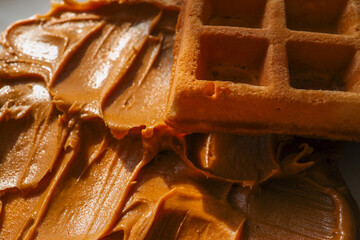 Close-up of a golden brown waffle resting on creamy peanut butter spread. The crispy texture of the waffle contrasts with the smooth, glossy surface of the peanut butter