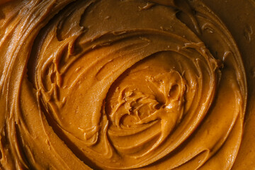 Close-up of creamy peanut butter spread with a rich, smooth texture. The golden-brown surface has visible swirls and ridges, showcasing its thick and glossy consistency