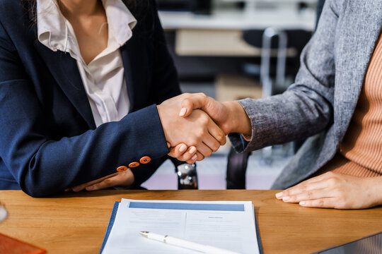 Two female lawyers in business suits shake hands at wooden desk, finalizing legal agreement. wooden gavel and scales represent justice as they discuss corporate law, compliance, dispute resolution.