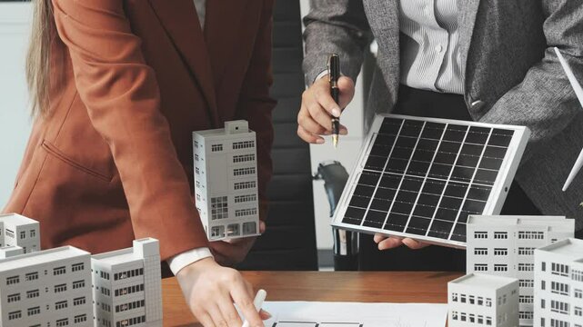Two businesswomen sit at a desk in a meeting, discussing property development while building a model of city tower. real estate investments, zoning laws, sustainable energy solutions. net zero