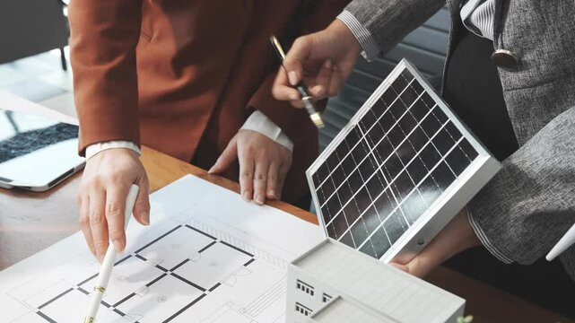 Two businesswomen sit at a desk in a meeting, discussing property development while building a model of city tower. real estate investments, zoning laws, sustainable energy solutions. net zero