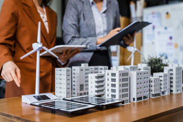 Two businesswomen sit at a desk in a meeting, discussing property development while building a model of city tower. real estate investments, zoning laws, sustainable energy solutions. net zero