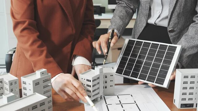 Two businesswomen sit at a desk in a meeting, discussing property development while building a model of city tower. real estate investments, zoning laws, sustainable energy solutions. net zero