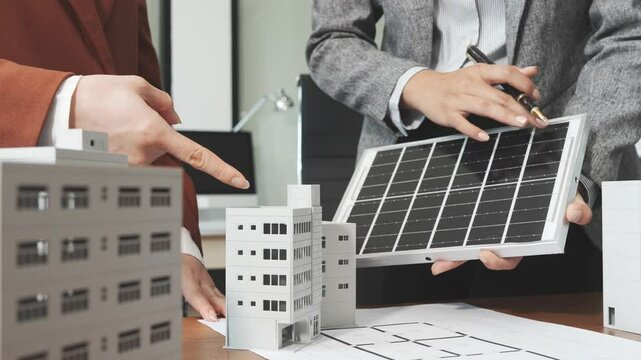 Two businesswomen sit at a desk in a meeting, discussing property development while building a model of city tower. real estate investments, zoning laws, sustainable energy solutions. net zero