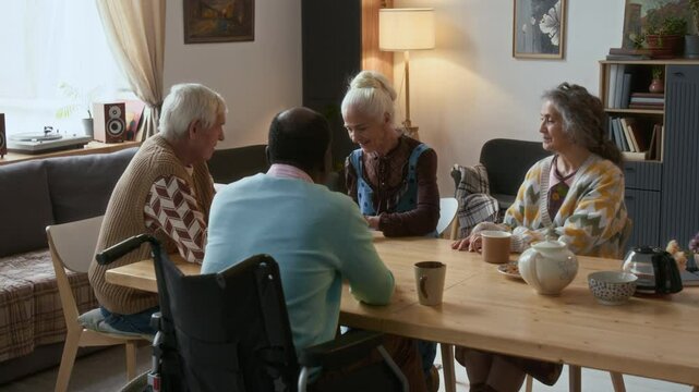 Multi-ethnic elderly friends sitting around table in cozy living room, playing cards and chatting while spending leisure time together in assisted living facility