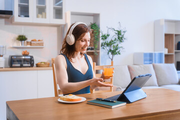 A white woman relaxes on the sofa in her living room, happily video calling her boyfriend. She enjoys a fresh salad with orange juice while using her laptop for a fun conversation.
