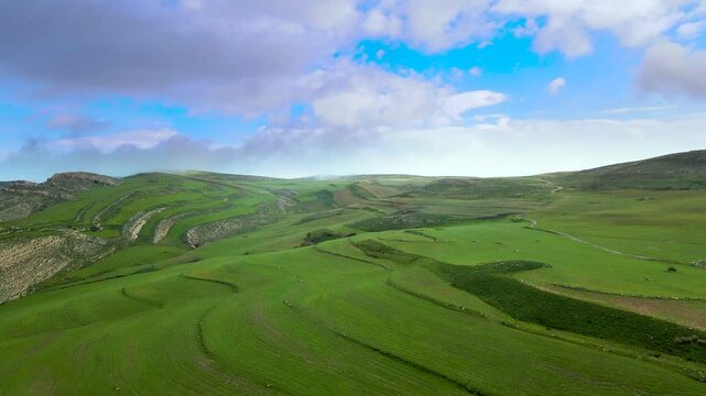 An aerial view of the scenic landscape in Djebba, Beja. The image showcases the vibrant green fields, rocky outcrops, and distant hills under a dynamic sky with clouds.