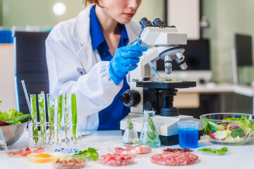 Fototapeta premium Middle-aged female laboratory worker wearing blue gloves conducts lab test on food ingredients lettuce, cucumber, pork. Using microscope, she analyzes nutrients, preservatives, food safety records.