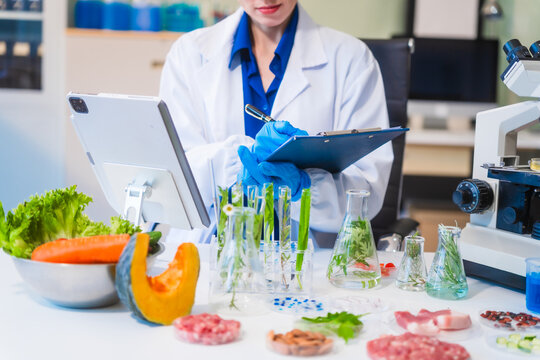 Middle-aged female laboratory worker wearing blue gloves conducts lab test on food ingredients lettuce, cucumber, pork. Using microscope, she analyzes nutrients, preservatives, food safety records.