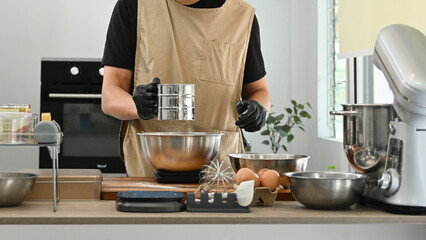 A man is making a cake and is using a sifter to sift flour. The kitchen is well-equipped with various bowls, spoons, and other utensils. The atmosphere is calm and focused.