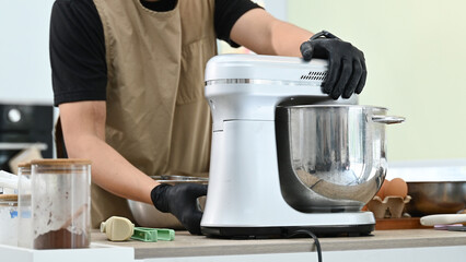A person is making a cake in a kitchen. The person is wearing a black apron and gloves. The cake batter is poured into a mixing bowl and then into a mixer.