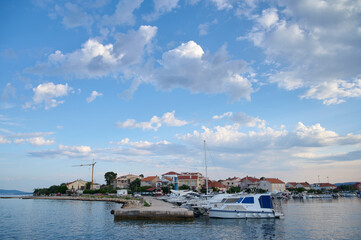 Fototapeta premium Calm harbor showcases various boats docked near a quaint coastal village under a blue sky with fluffy clouds.