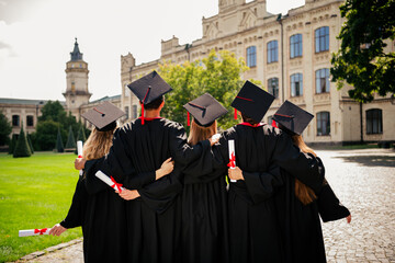 Rear portrait of group students hug hold diploma mortarboard gown graduate college building outdoors