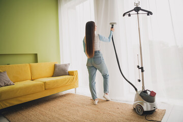 Fototapeta premium Young Woman Using Steam Cleaner on Curtain in Stylish Modern Interior with Comfortable Couch and Daylight