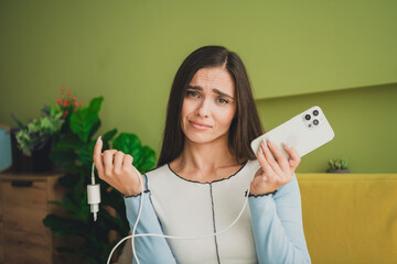 Young brunette woman displaying a disconnected phone charger with a humorous expression in a cozy...