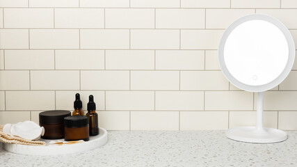 Skincare products and mirror with LED light on marble bathroom table top, tile wall. Cream jar, lotion and serum brown bottles on dressing table