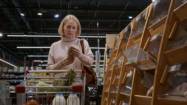 Front view shot of blonde adult woman choosing fresh bread in bakery at supermarket and putting it in cart