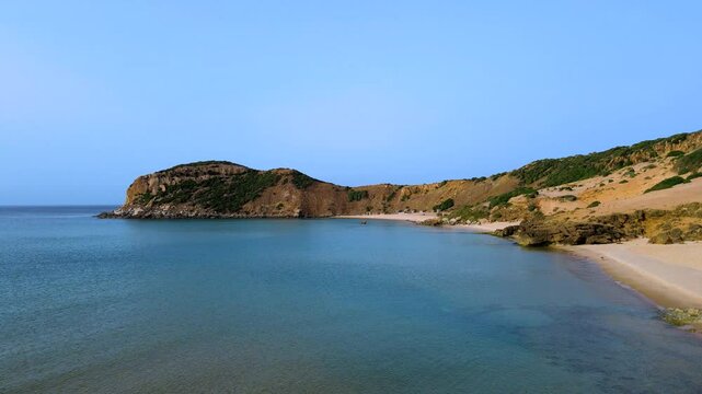 A serene beach scene at El Gliaa in Bizerte, featuring calm turquoise waters and a sandy shoreline. The rocky hill in the background adds depth and character to this tranquil setting.