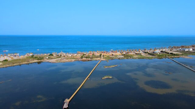 This video captures an expansive aerial view of Sidi Ali Makki Beach in Bizerte.The long wooden pier stretches across the water, connecting to a vibrant beach area filled with straw huts and buildings