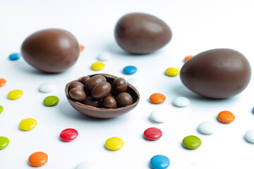 Chocolate Easter eggs with a broken one revealing chocolate-covered treats inside, surrounded by multicolored candy pieces on a white background. Ideal for festive branding, confectionery advertising.