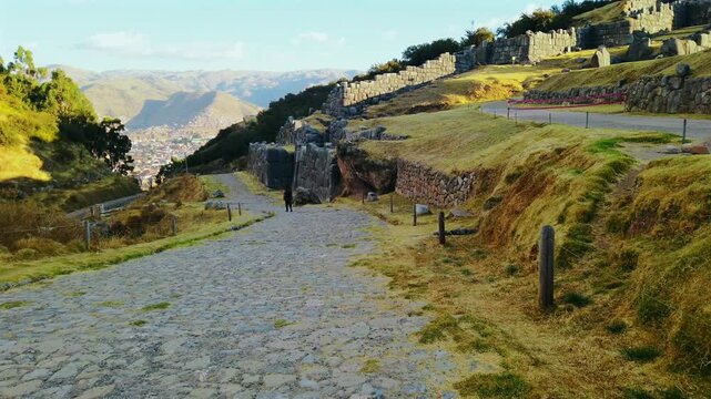 Unique view of Sacsayhuaman ruins with a solo traveler walking on the ancient Inca road. Follow shot overlooking the ruins and the city of Cusco in the valley