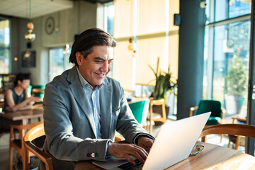 Smiling businessman drinking coffee while working on laptop in modern cafe