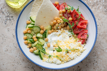 Savory yogurt bowl with cucumber, tomatoes, chickpeas and grated egg, horizontal shot on a beige granite surface, middle closeup