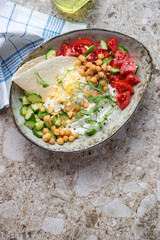 Yogurt bowl with vegetables, chickpeas, grated egg and flat bread, top view on a beige granite background, vertical shot with space