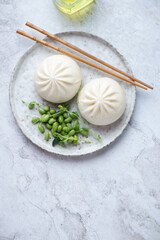 Plate with freshly cooked chinese baozi and edamame beans, vertical shot on a white stone background with space, flat lay