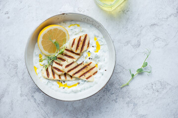 Bowl with greek tzatziki sauce and grilled halloumi, horizontal shot on a white stone background with space, view from above