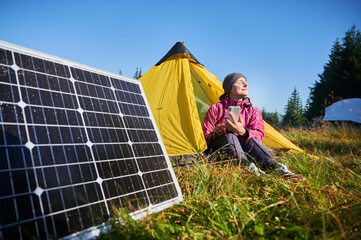 Happy woman sits in front of yellow tent, charging her phone by using solar panel, highlighting use...