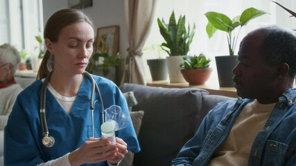 Tilt up shot of Caucasian female doctor in scrubs explaining how to use inhaler to elderly African American patient, providing medical guidance and support in nursing home