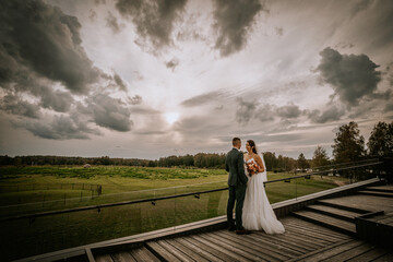 A newlywed couple stands on a wooden terrace, gazing at each other against a dramatic cloudy sky,...