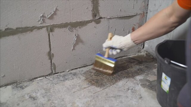 Construction worker applies waterproofing membrane with a brush on a concrete floor next to a cinder block wall
