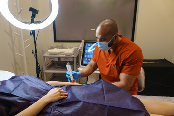Doctor wearing orange uniform and face mask using laser device for tattoo removal from patient's...