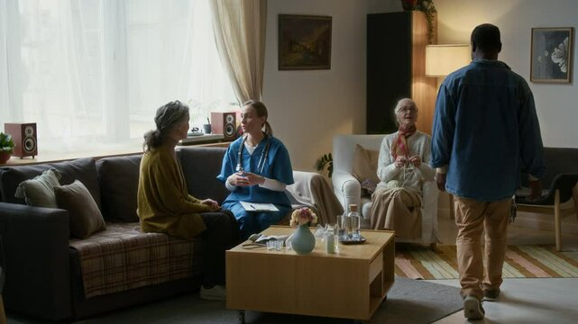 Nurse sitting on couch, talking to elderly woman and taking notes while another seniors talking nearby in cozy living room of assisted living facility