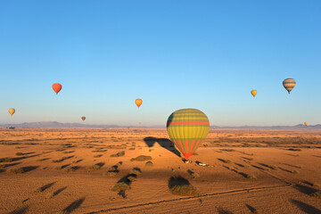 Hot air balloons float over a vast desert landscape at sunrise, casting long shadows on the golden sand. The clear sky and distant mountains add depth.