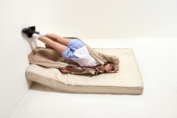 Naklejka premium Young woman lying on mattress with legs propped against wall, dressed in casual yet business-inspired attire. Concept of burnout, unconventional relaxation, surreal office escape.