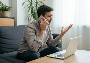 Frustrated young adult man on phone call sitting with laptop in modern living room