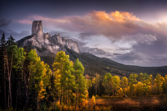 View of chimney rock at sunset surrounded by colorful autumn forest and dramatic sky, Cimarron, Colorado, United States.