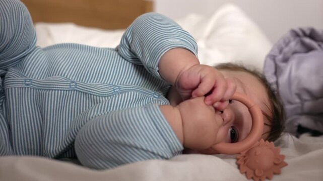 Baby lying on bed, holding teething ring, exploring object with mouth, early sensory development, chewing toy, soothing gums, infant play, calming moment, home setting