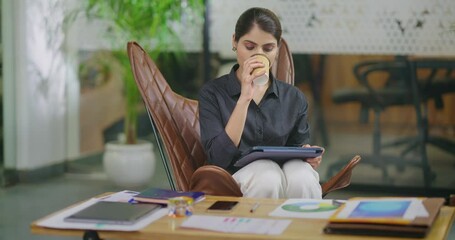 Indian young adult gen z busy woman lady use tablet device look screen do online data check email work girl female intern staff pick up paper cup drink tea coffee have day break time job office place