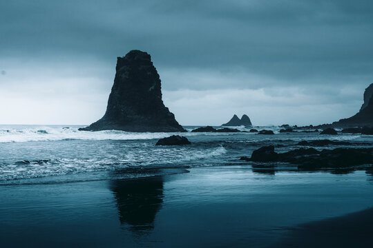 View of dramatic sea stacks and rugged rock formations with crashing waves under a cloudy dark sky, Playa de Benijo, Santa Cruz de Tenerife, Spain.