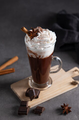Hot chocolate with whipped cream, cinnamon and spices in a tall glass on a dark background.