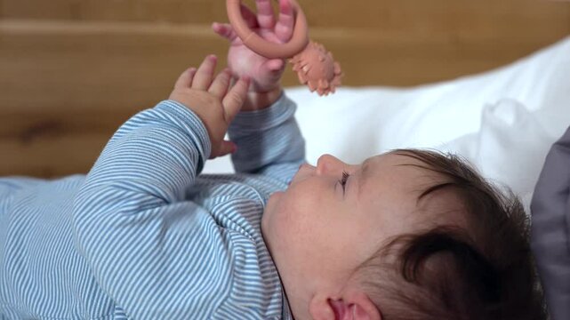 Baby lying on soft bed holding teething ring with hedgehog design, gazing upward with curiosity, gripping toy firmly, engaging in sensory play and early motor skill development