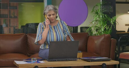 Indian asian older woman lady sitting on sofa couch use laptop looking screen do online work check bills e-bank data app serious senior adult female boss worker talking on phone call at office - Powered by Adobe