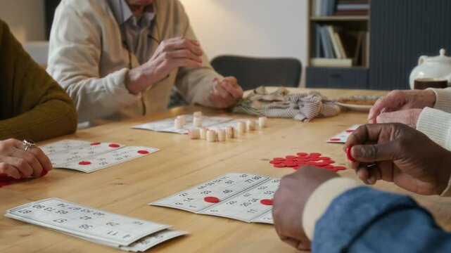 Elderly man playing bingo with company of friends, using tokens and cards at wooden table in cozy living room of retirement home
