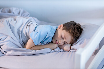 A young boy wearing a blue shirt is peacefully sleeping on his cozy bed tucked in with soft blankets and surrounded by comfortable linens.