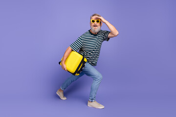 Smiling Senior Man Carrying a Yellow Suitcase with a Playful Pose on a Purple Background
