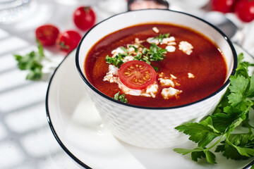 Spanish Cold Tomato Gazpacho Soup garnished with parsley, feta cheese and spices with toasted bread slices, tomatoes and herbs on white table